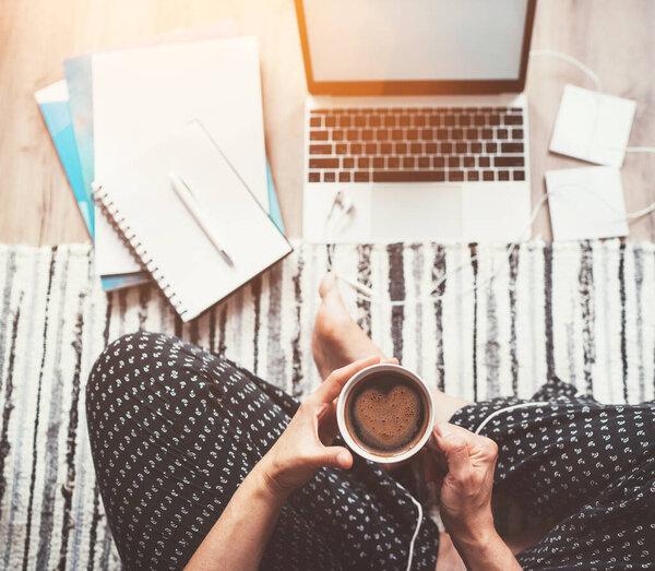  Business woman dressed pajamas enjoying morning coffee with heart shape foam sitting on living room floor office with laptop, papers and other stuff.Distance work in worldwide quarantine time concept