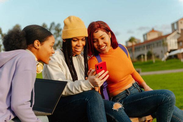 multiracial group of 3 female friends sitting on a bench on a university campus using a cell phone and a laptop. classmates having a good time during a break