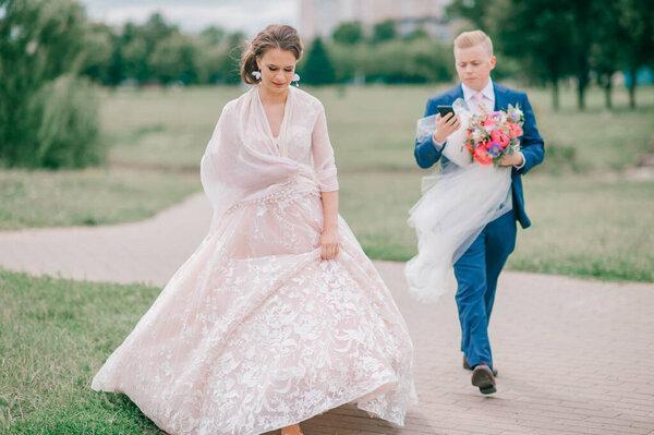 Groom carrying brides veil and talking by smartphone. Wedding couple walking outdoor.