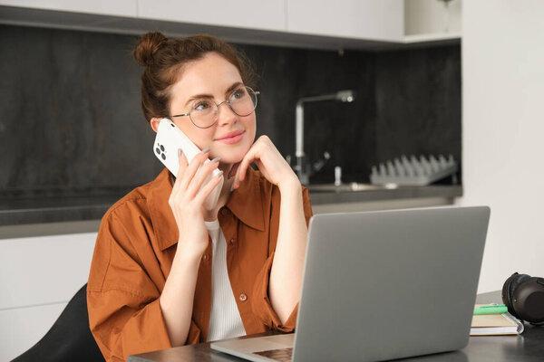 Portrait of young self-employed woman, entrepreneur working from home, freelancer calling client. Girl making an order, talking to someone on phone, sitting in kitchen with laptop.