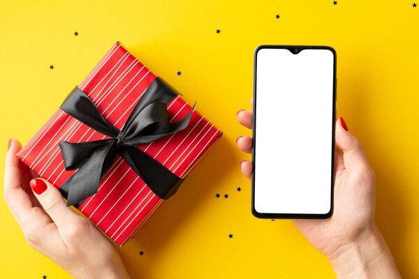 Black friday shopping concept. First person top view photo of woman's hands holding smartphone and big red giftbox with ribbon bow over confetti on isolated yellow background with empty space