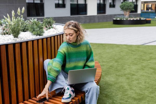 young woman in green sweater sitting on wooden bench with laptop while taking smartphone with blank screen 