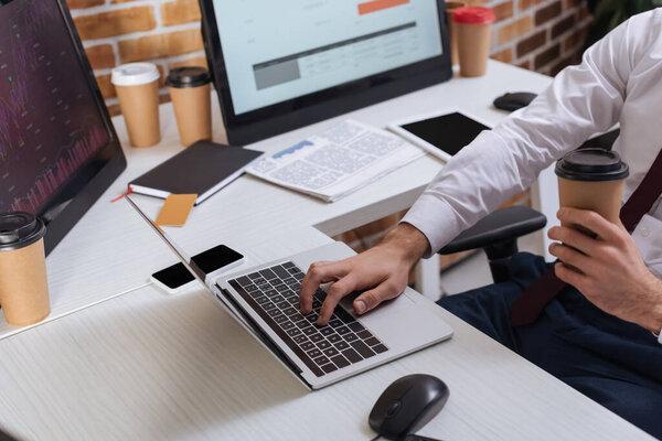 Cropped view of businessman holding coffee to go and checking financial stocks on laptop in office 