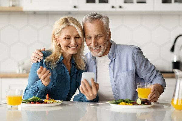 Portrait Of Happy Senior Spouses Using Smartphone During Breakfast In Kitchen, Married Elderly Couple Making Video Call On Mobile Phone Or Checking New App, Closeup Shot With Free Space