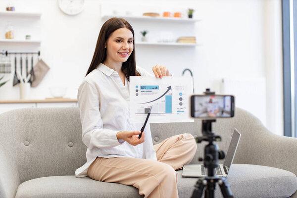 Concept of technology and blogging. Young caucasian brunette financier woman filming herself on cell phone which fixed on tripod and showing financial statistics to subscribers in living room.