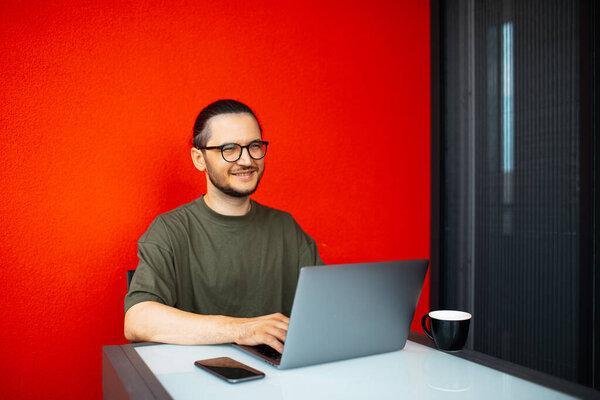 Portrait of young smiling man working at laptop on background of colourful red wall. Business people. Wearing eyeglasses and sweater. Typing on keyboard.