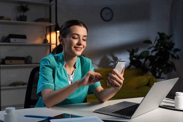 smiling nurse in uniform sitting at table and using smartphone during night shift