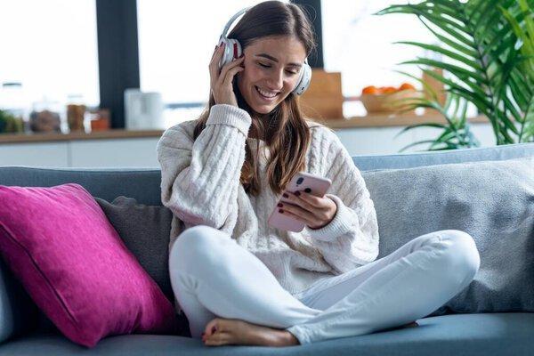 Shot of smiling young woman listening to music with smartphone while sitting on couch at home.