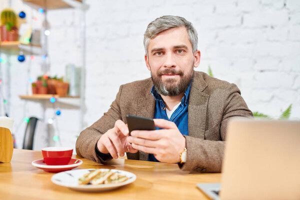 Young serious man in formalwear sitting by table in front of camera, having tea with snack and messaging in smartphone