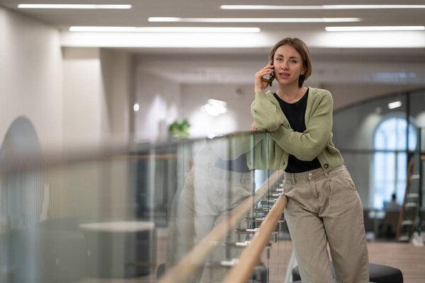 Successful self-sufficient woman talking on phone standing inside business center building calling taxi or courier. Young businesswoman leans on glass partition making call to business partners 