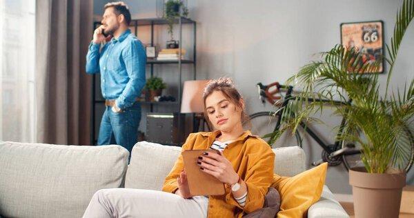Portrait of happy beautiful young Caucasian woman sitting on sofa in modern living room in apartment typing on tablet while her husband speaking on smartphone behind, family couple, marriage concept