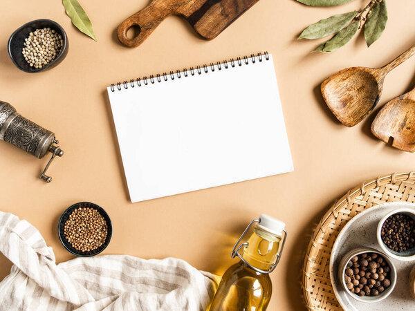 Kitchen flat lay with open notebook for culinary text and small bowls various dry spices, wood kitchen utensils, olive oil in glass bottle on a beige background. Top view. copy space
