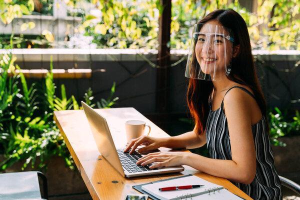 Young 20s Asian woman looking at camera while wearing a protective mask with laptop, mobile phone, and notebook for working outside in casual business look. Corona Virus - Covid 19 work online concept