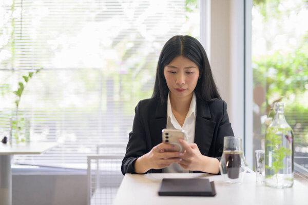 一位专业的亚洲女商人在明亮、现代化的咖啡馆里，一边喝着咖啡，一边在绿植旁使用她的智能手机工作。