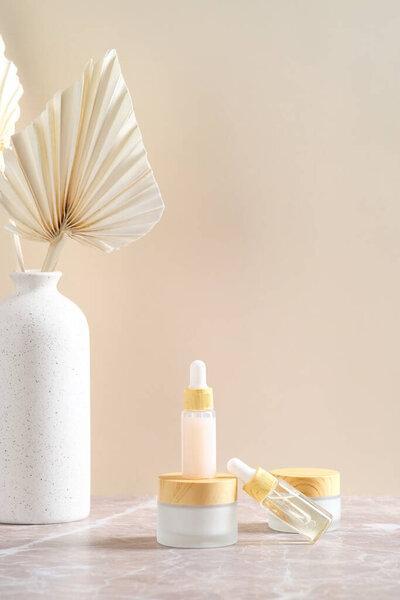 Cosmetic dropper bottles with essential oils, body cream and vase of dry flowers on beige background.