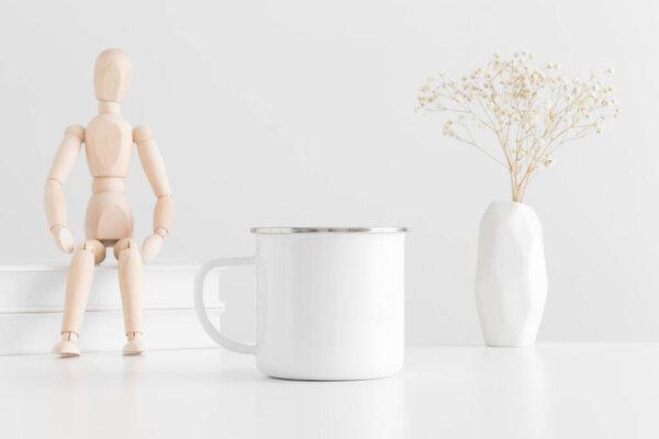 Enamel mug mockup with books, manikin and a gypsophila in a vase on a white table.