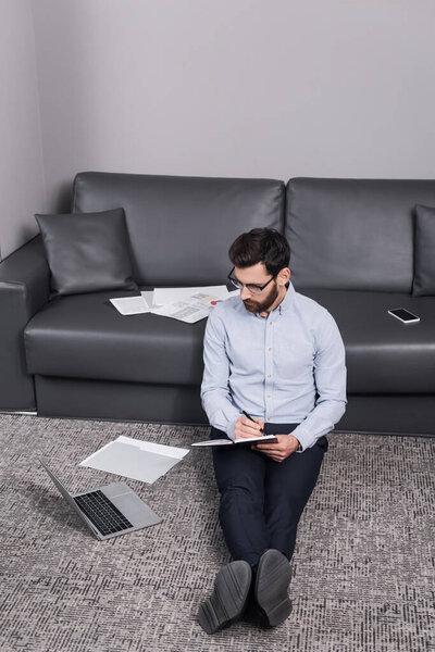 bearded freelancer in eyeglasses sitting on carpet and writing in notebook near laptop and couch 