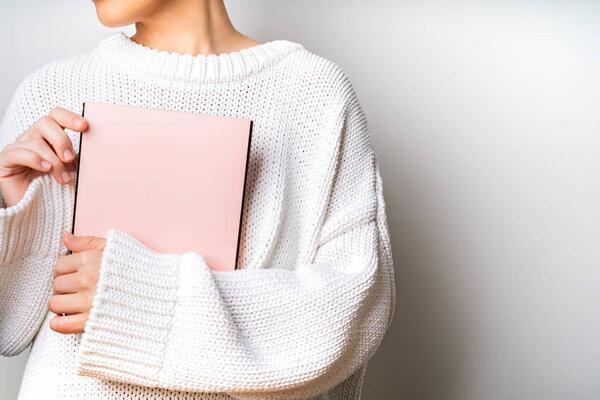 Close view of woman in white woolen sweater holding a book with empty pink cover in hands. Free space for your mock up of reading book concept background.
