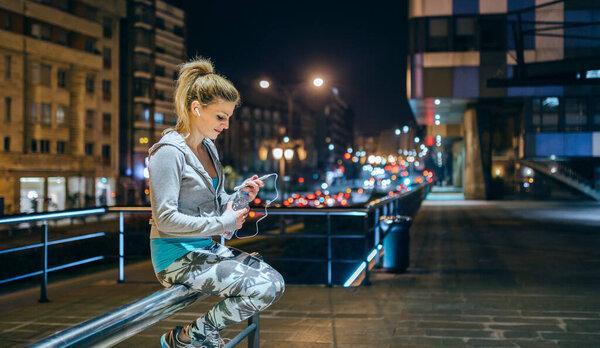 Smiling woman looking her coach messages on mobile app while open bottle of water after training. Female young runner with earphones resting sitting over banister at night on town. Right copy space.