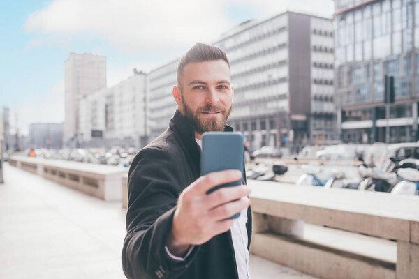 Young bearded caucasian man outdoor using smartphone taking selfie smiling outdoor