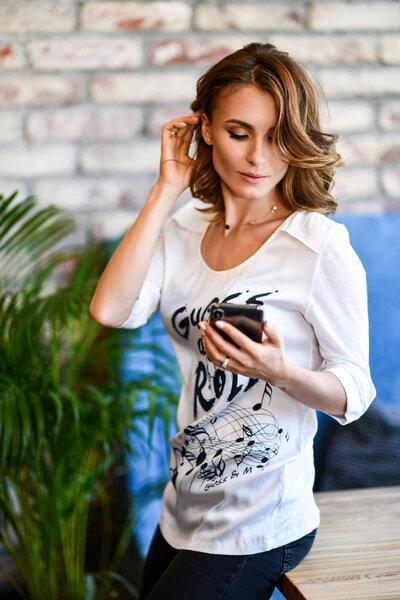 young brunette in a white T-shirt is standing near a table in a cafe with a phone in her hands, she is waiting for her friend or colleague to meet. Not a forced environment. Modern interior.