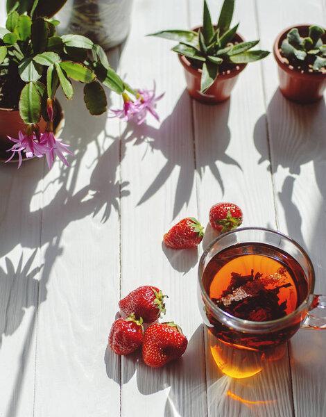 Close-up image of glass cup of tea, red strawberries, flowers in pots on a light wooden background.