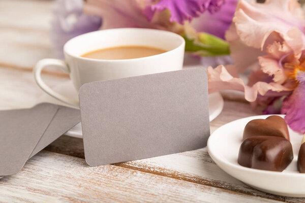 Gray business card with cup of cioffee, chocolate candies and iris flowers on white wooden background. side view, copy space, still life. Breakfast, morning, spring concept.
