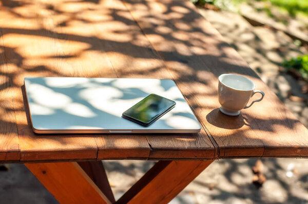 View on a closed laptop pc and a smartphone coffee mug on the table in the garden in a home office or home school enviroment on a sunny day.