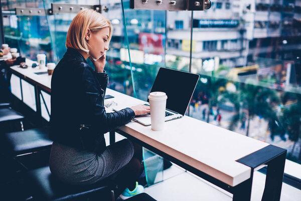 Stylish businesswoman with short haircut talking on smartphone and booking tickets while working at modern netbook with mock up area connected to wireless 4G internet in coffee shop with cityscape