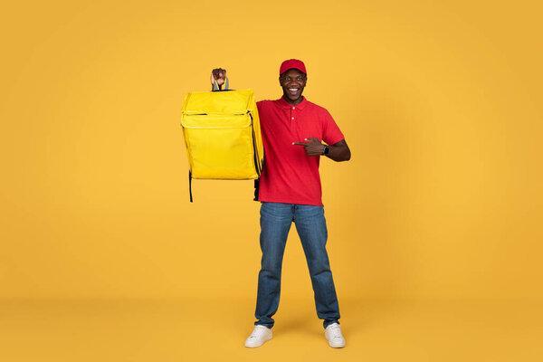 Smiling young african american delivery man in uniform point finger at big backpack, isolated on orange studio background. Delivery service recommendation, ad and offer, work