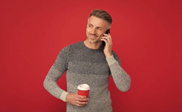concept of communication. smiling grizzled guy speaking on phone on red background. hoary man drinking coffee. male fashion model hold cup and smartphone. morning coffee.