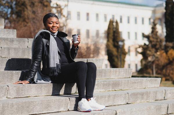Portrait of a smiling young African American girl with pigtails with coffee walking in the street on a sunny day. Outdoor photo.