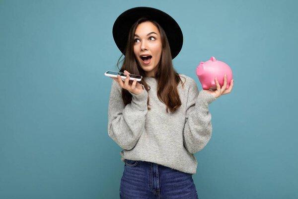 Portrait photo of happy positive sincere young charming winsome woman with dark hair wearing casual gray sweater and black hat isolated on blue background with copy space, holds pink pig money box and