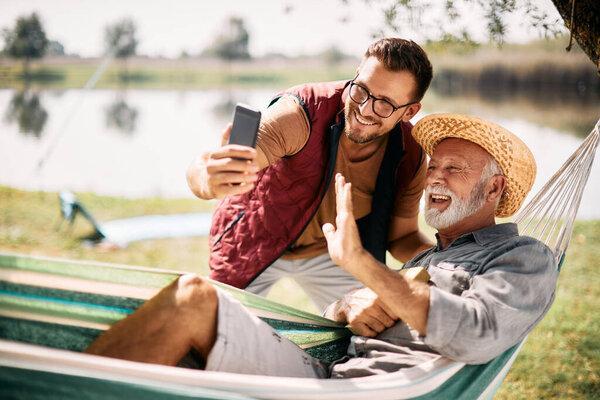 Happy senior man and his son greeting someone via video call while enjoying a day in nature.