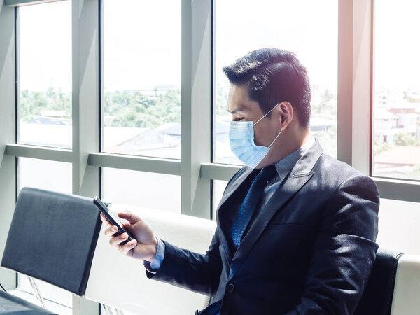 Asian businessman in suit wearing protective face mask using mobile phone while sitting in modern office building near huge glass window.