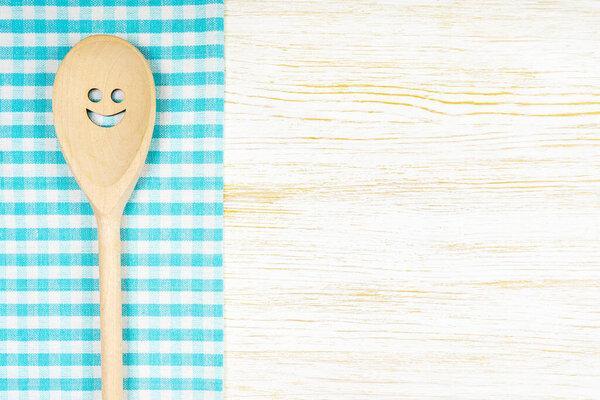 Wooden spoon with smile on blue checkered tablecloth on white wooden surface. Mockup for menu or recipe, restaurant, website with cooking. Kitchen food background, template, flat lay with copy space