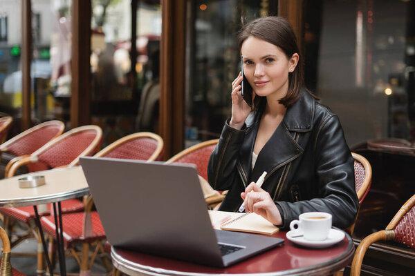 young freelancer in black jacket talking on smartphone near laptop and cup of coffee on table in outdoor cafe in paris