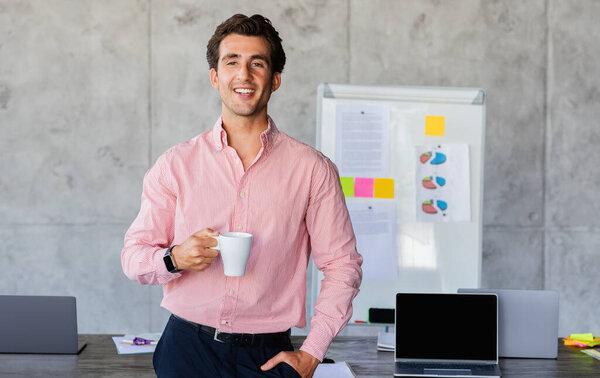 Happy handsome young businessman wearing formal outfit standing by workplace at office, drinking coffee and smiling after successful business meeting, have break, copy space