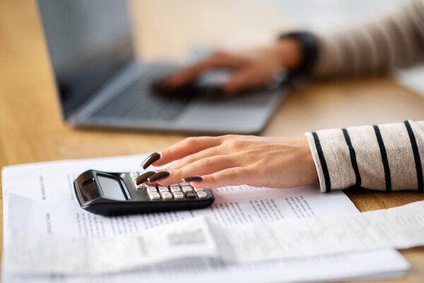 Focused middle eastern millennial woman calculating finances with a calculator and laptop at a wooden desk, surrounded by documents and displaying a close-up of her hand