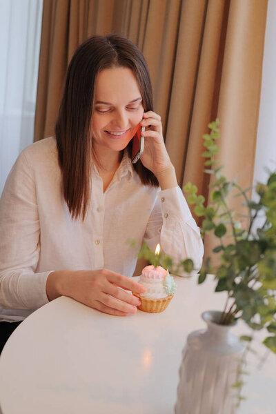 Beautiful cute smiling woman talking on mobile phone and smiling happily while looking at festive dessert holding cake with candle expressing happiness