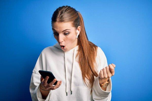 Young redhead sportswoman doing sport listening to music using earphones and smartphone Pointing down with fingers showing advertisement, surprised face and open mouth
