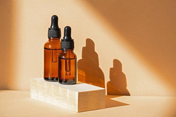 Improvised 3d wooden podium with two glass dropper bottles on brown background, in rays of sunlight. Hard shadow from objects falls on surface. Concept of the beauty industry.