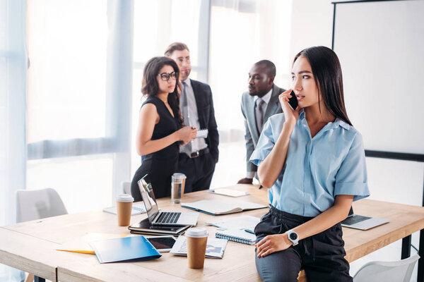 selective focus of asian businesswoman talking on smartphone and colleagues behind in office