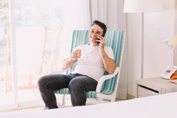 Young man sitting in a rocking chair in his bedroom, talking on the mobile and having a coffee, with the light coming through the window. Concept of working, vacation, connection and smartphone.