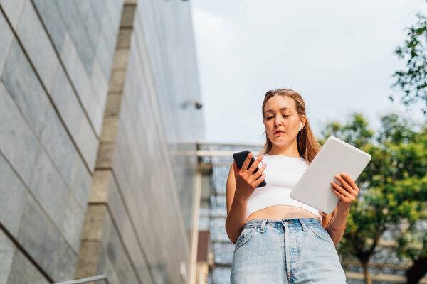 Contemporary caucasian multitasking business woman outdoor using tablet calling with smartphone and wireless earphones