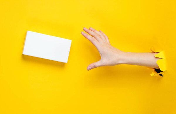 Female hand takes white box through torn yellow paper. Minimalistic creative fashion concept