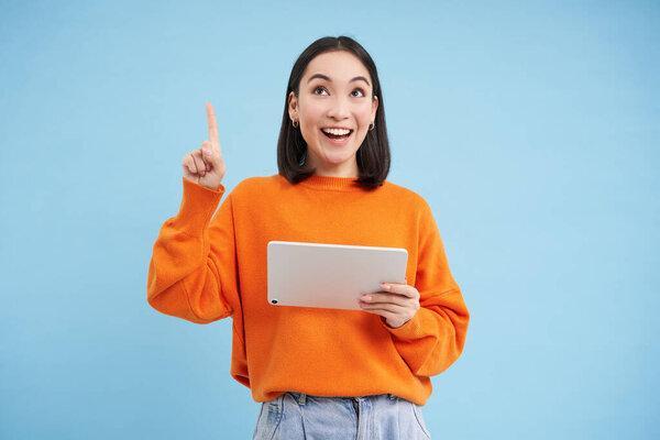 兴奋的韩国女人，带着数码平板电脑，指着空旷的空间，广告横幅，手持小玩艺儿，展示弹药，蓝色背景.