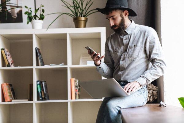 Photo of thinking young man wearing hat using laptop and cellphone while sitting on table in cafe indoors