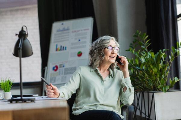 Smiling businesswoman holding pen while taking on smartphone near plants and blurred flip chart in office 