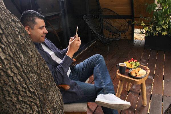 Dark-haired 40-year-old Latino adult man uses his cell phone for video calls and messages while having breakfast in a cabin in the middle of the woods